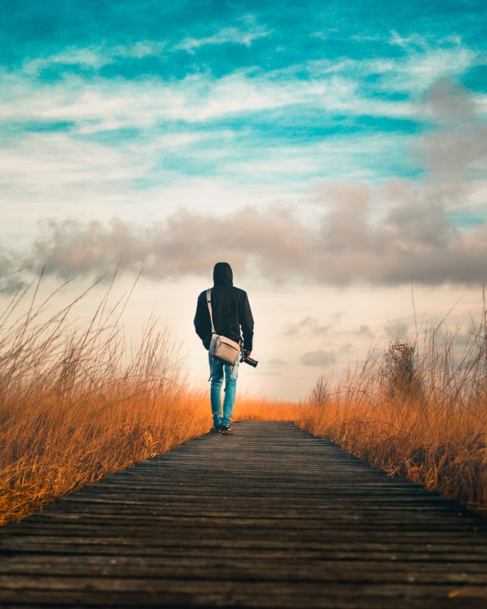 Person in Black Hoodie Jacket Walking on Wooden Boardwalk Between Brown Grass